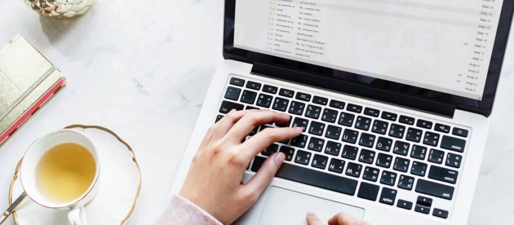 Woman's hands typing on a laptop keyboard with a teacup of tea to her left.