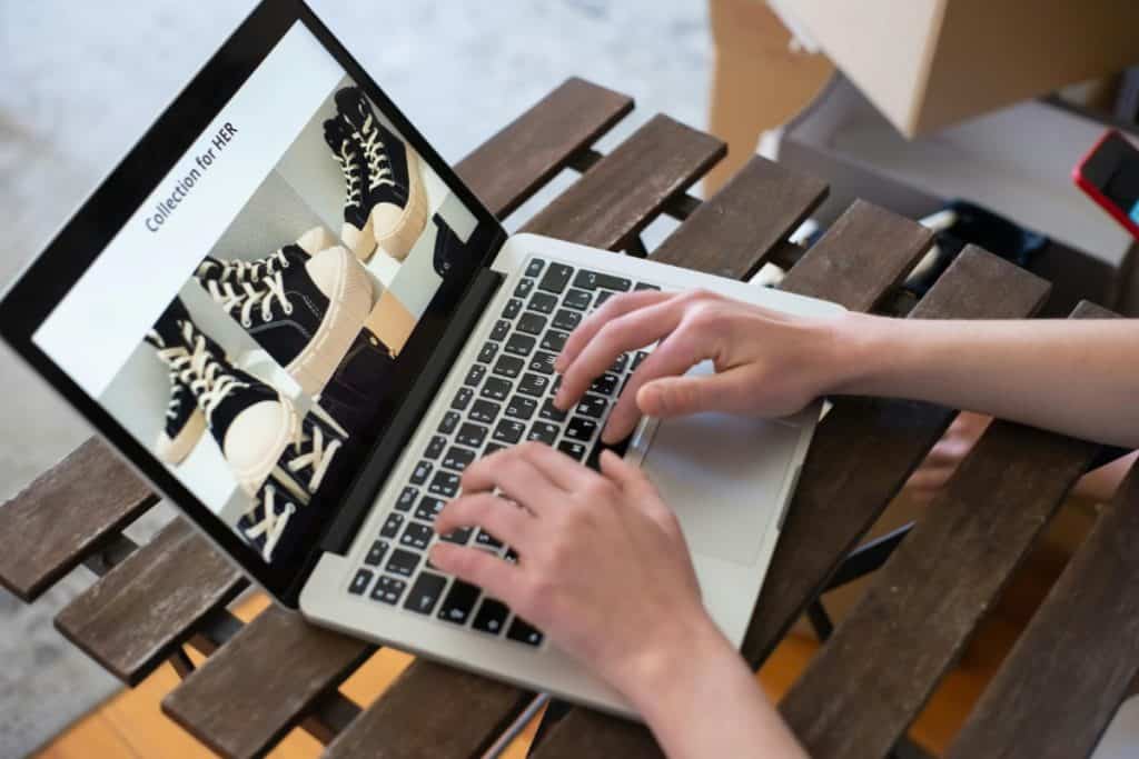 Hands typing on a laptop displaying sneakers products, outdoors at a garden table.